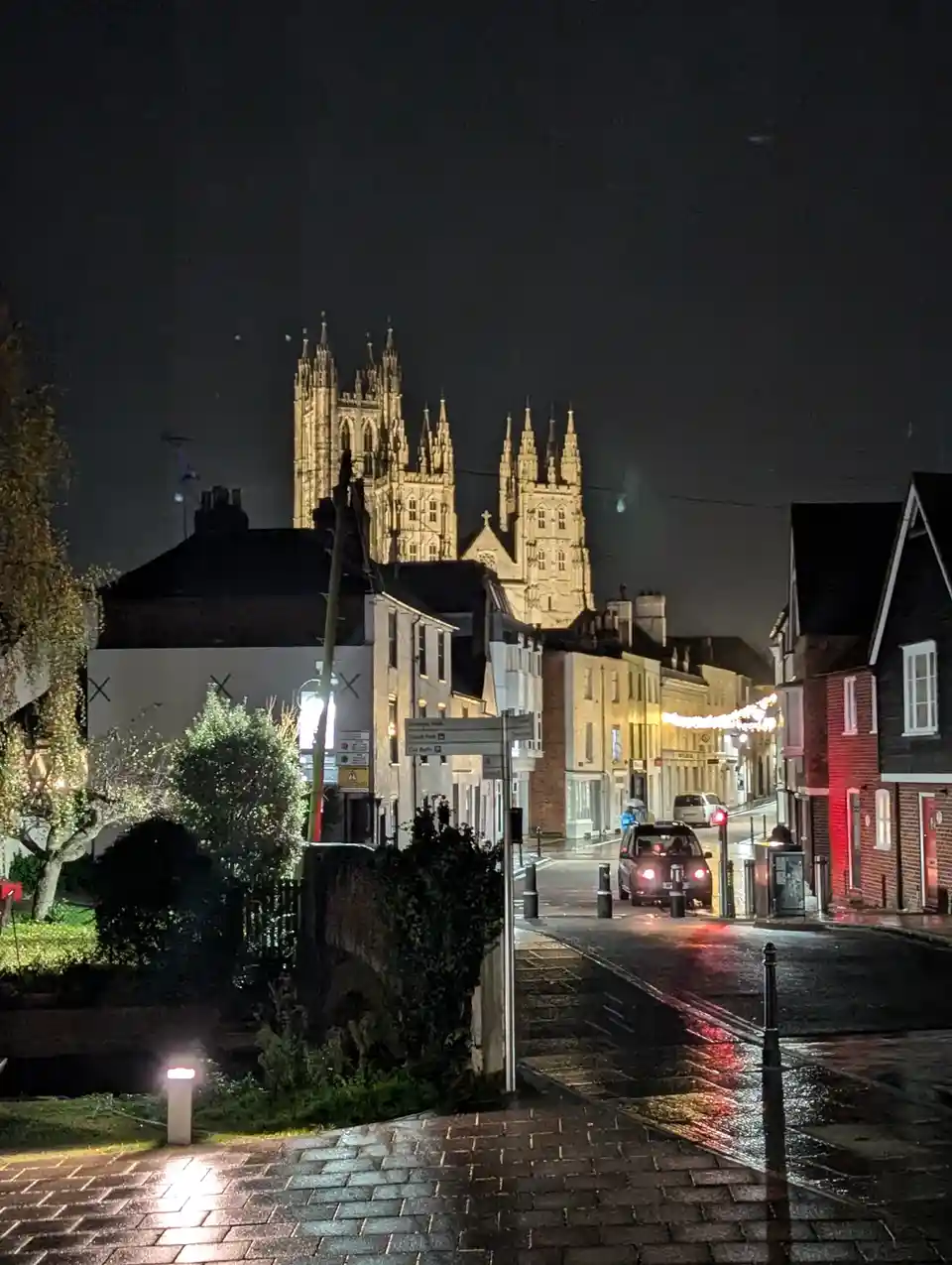 A nighttime street scene in a town with a large, illuminated gothic cathedral in the background, wet pavement reflecting…