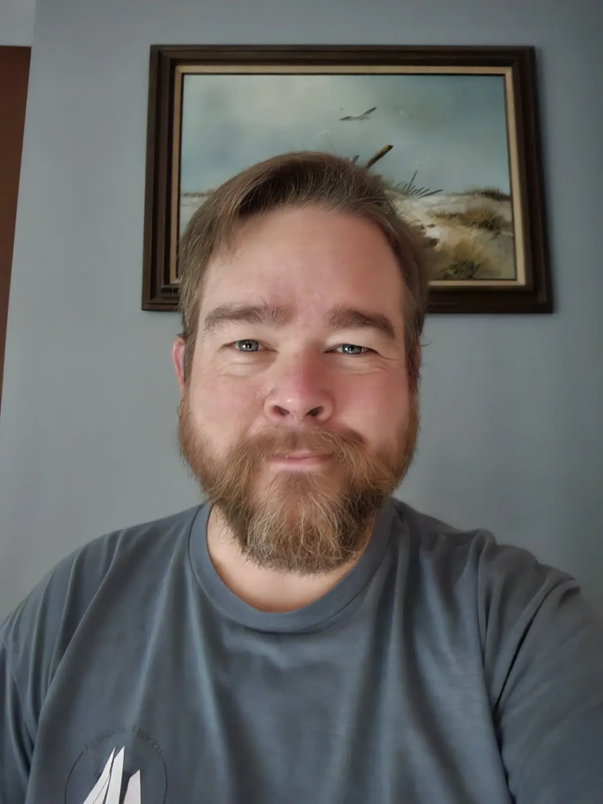 A man with a beard and blue eyes looks at the camera with a slight smirk while standing in front of a framed painting of a…