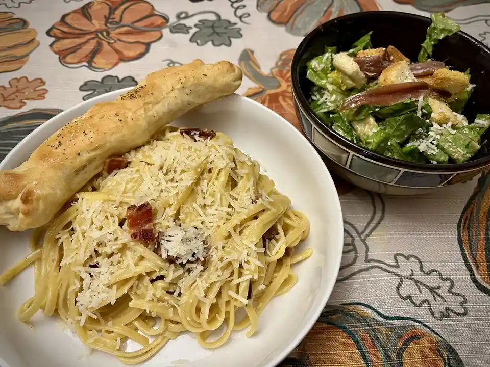 A plate of creamy pasta carbonara topped with grated cheese and crispy bacon, accompanied by a breadstick, sits next to a…