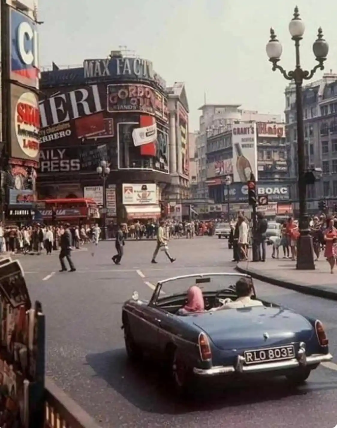 A vintage photograph of a busy London street corner, likely Piccadilly Circus, filled with pedestrians, classic cars, and…
