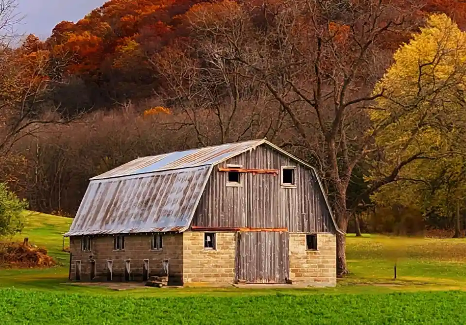 A rustic, weathered barn with a corrugated metal roof and stone foundation sits in a vibrant green field, surrounded by…