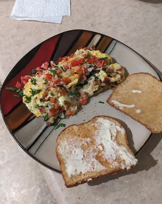 A plate of breakfast food, including a large, loaded scrambled egg dish with tomatoes, cheese, and herbs, alongside two…