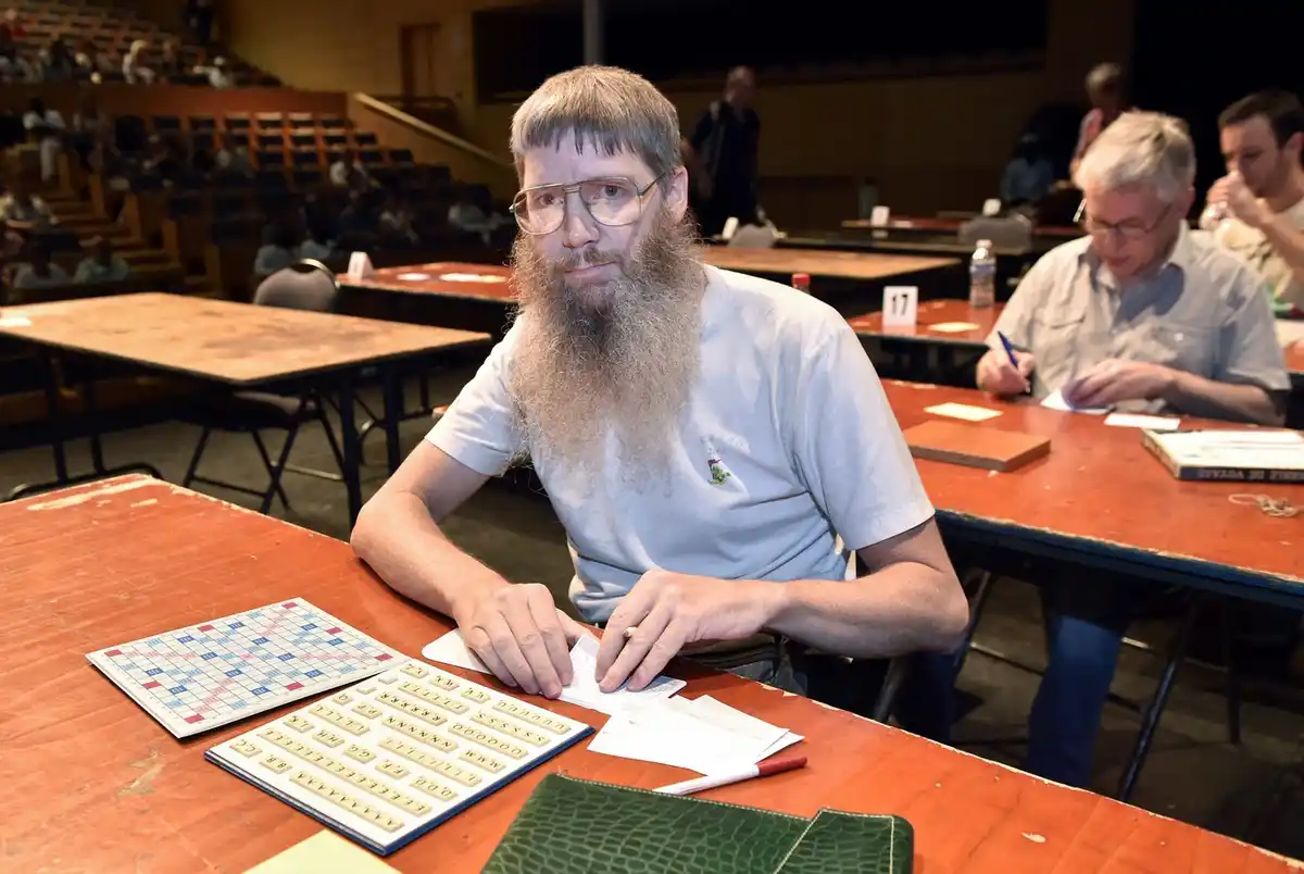 a man with a long grey beard and glasses sits at a wooden table in what appears to be a Scrabble tournament, looking…