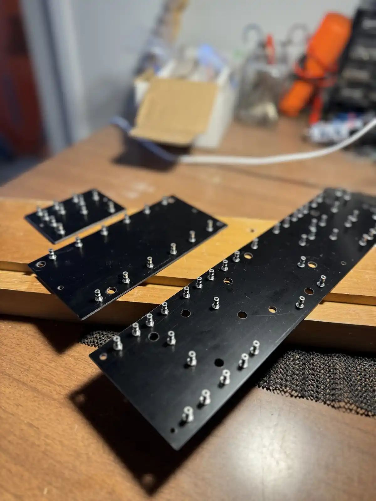 Three black keyboard plate pieces with numerous silver screws and drilled holes lie on wooden supports on a cluttered…