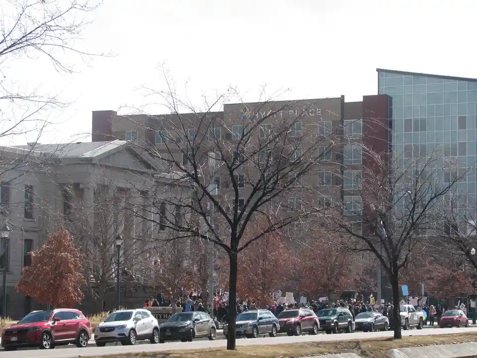 A street view shows a crowd of protesters holding signs behind a line of parked cars, with a large Hyatt Place hotel visible…