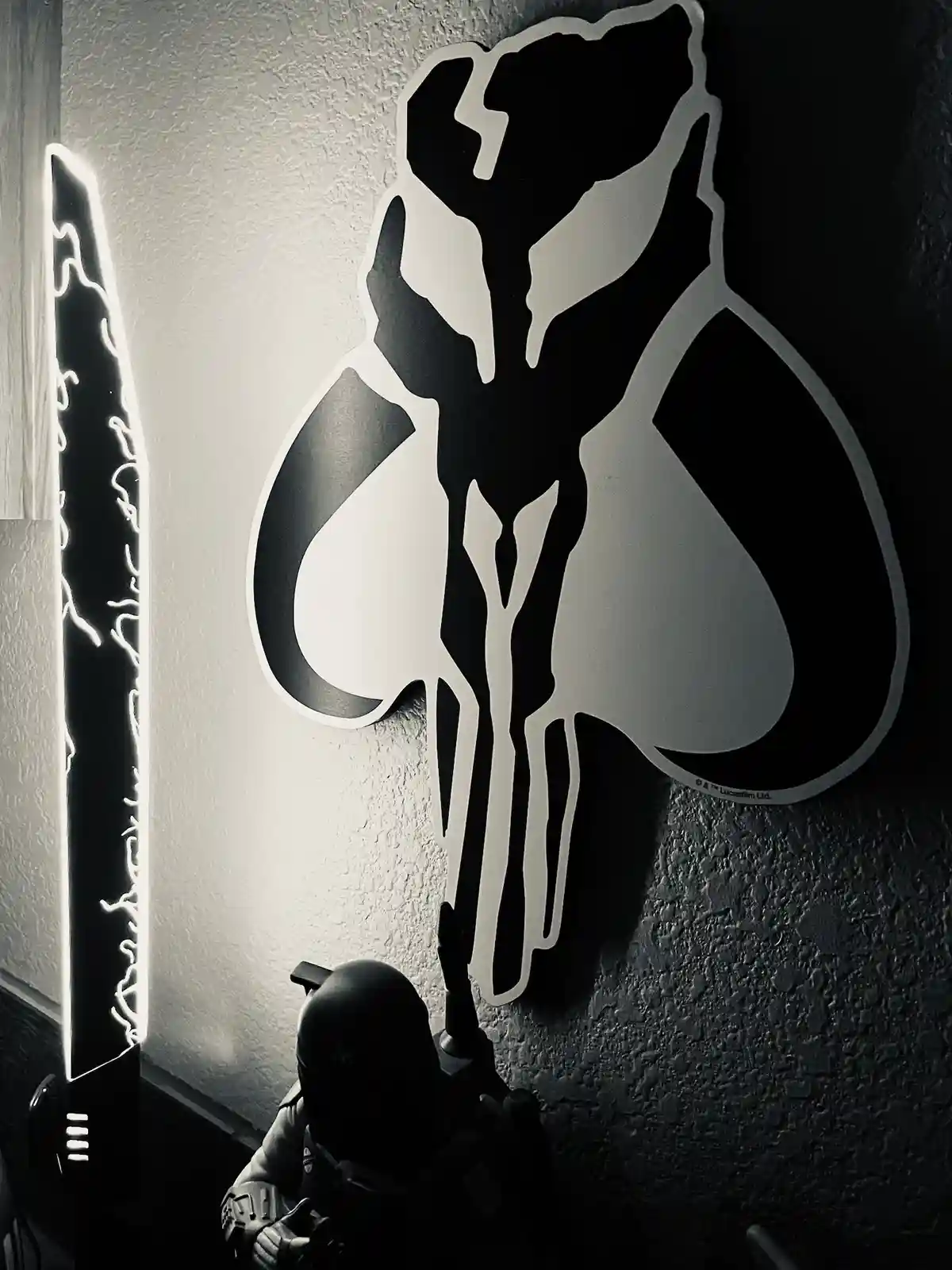 A black-and-white photo shows a person in a Mandalorian helmet and armor standing in a dimly lit room, looking up at a…