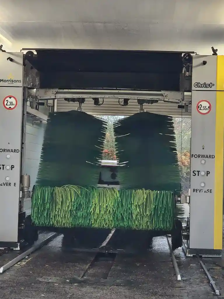 A car is being washed in an automatic car wash, with large green brushes and water spraying. The car's rear window reflects…