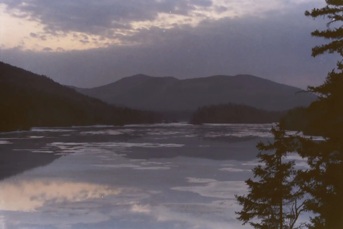 A serene, low-light photograph of a partially frozen lake with mountains in the background and evergreen trees framing the…