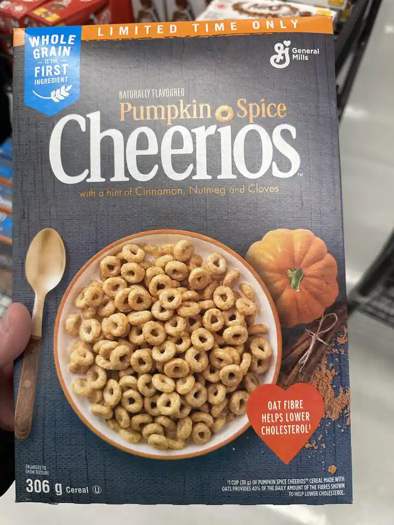 A person holds a box of General Mills Pumpkin Spice Cheerios cereal in a store aisle. The box features a bowl of cereal, a…