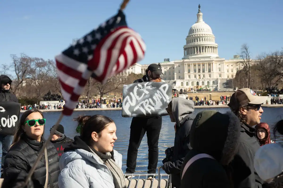 A man with a beard and black cap holds a handmade sign reading 'FUCK TRUMP' in white graffiti-style letters, standing near…