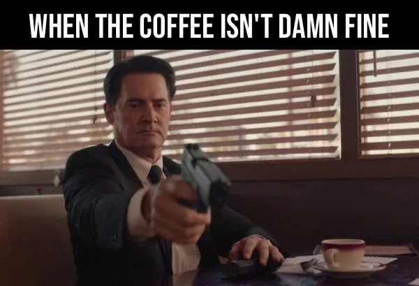 A man in a suit sits at a diner table, holding a handgun pointed directly at the viewer, with a coffee cup on the table…