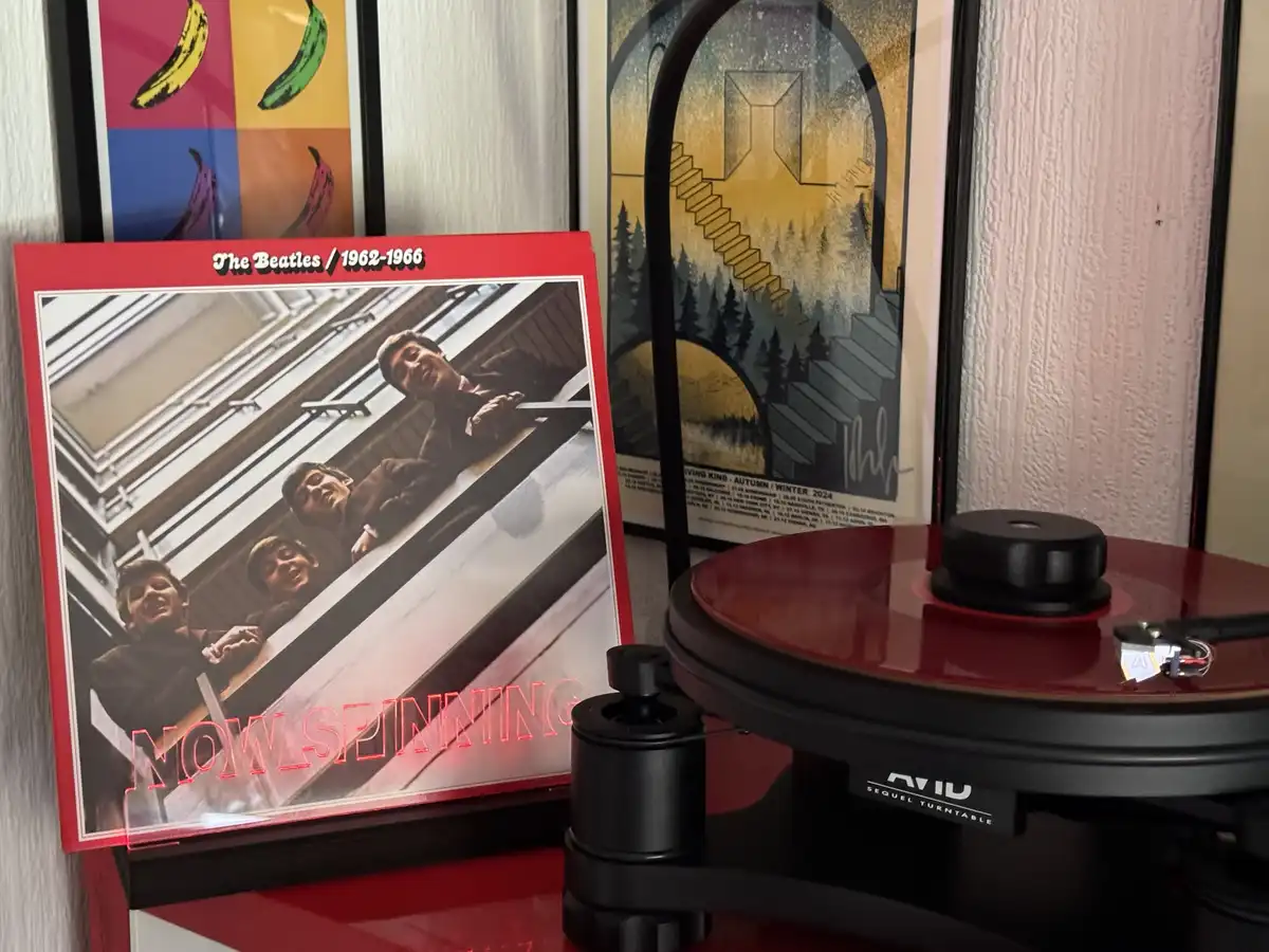 A red vinyl record player sits next to a framed album cover of The Beatles' '1962-1966' compilation. The album cover…