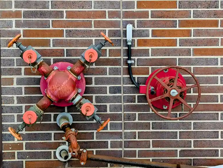 A red fire hydrant with multiple orange valves and chains is mounted on a brick wall, next to a red fire alarm pull station…