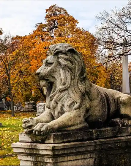 A weathered stone lion statue lies resting on a pedestal in a cemetery, surrounded by autumn trees with orange leaves and…