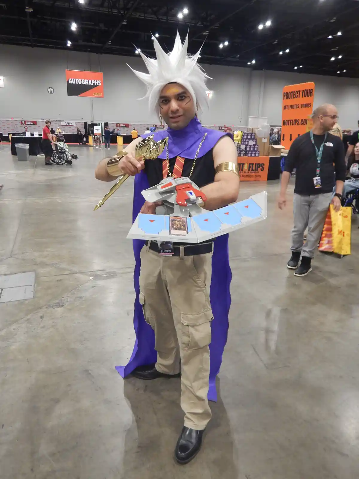 A man in a cosplay of a Yu-Gi-Oh! character with white spiky hair, purple cape, and gold accessories stands in a convention…