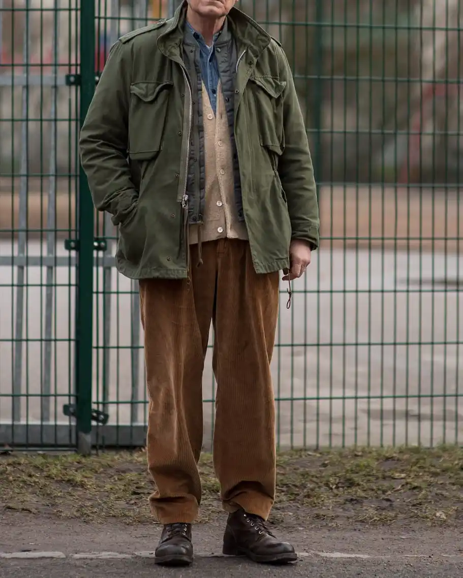 a man stands outdoors in front of a green metal fence, wearing a layered outfit including an olive green jacket, a beige…