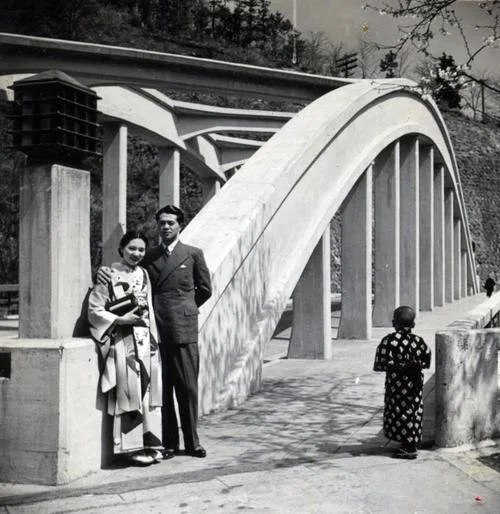 A black-and-white photo shows a man and woman in traditional Japanese attire standing near a large, modern concrete arch…