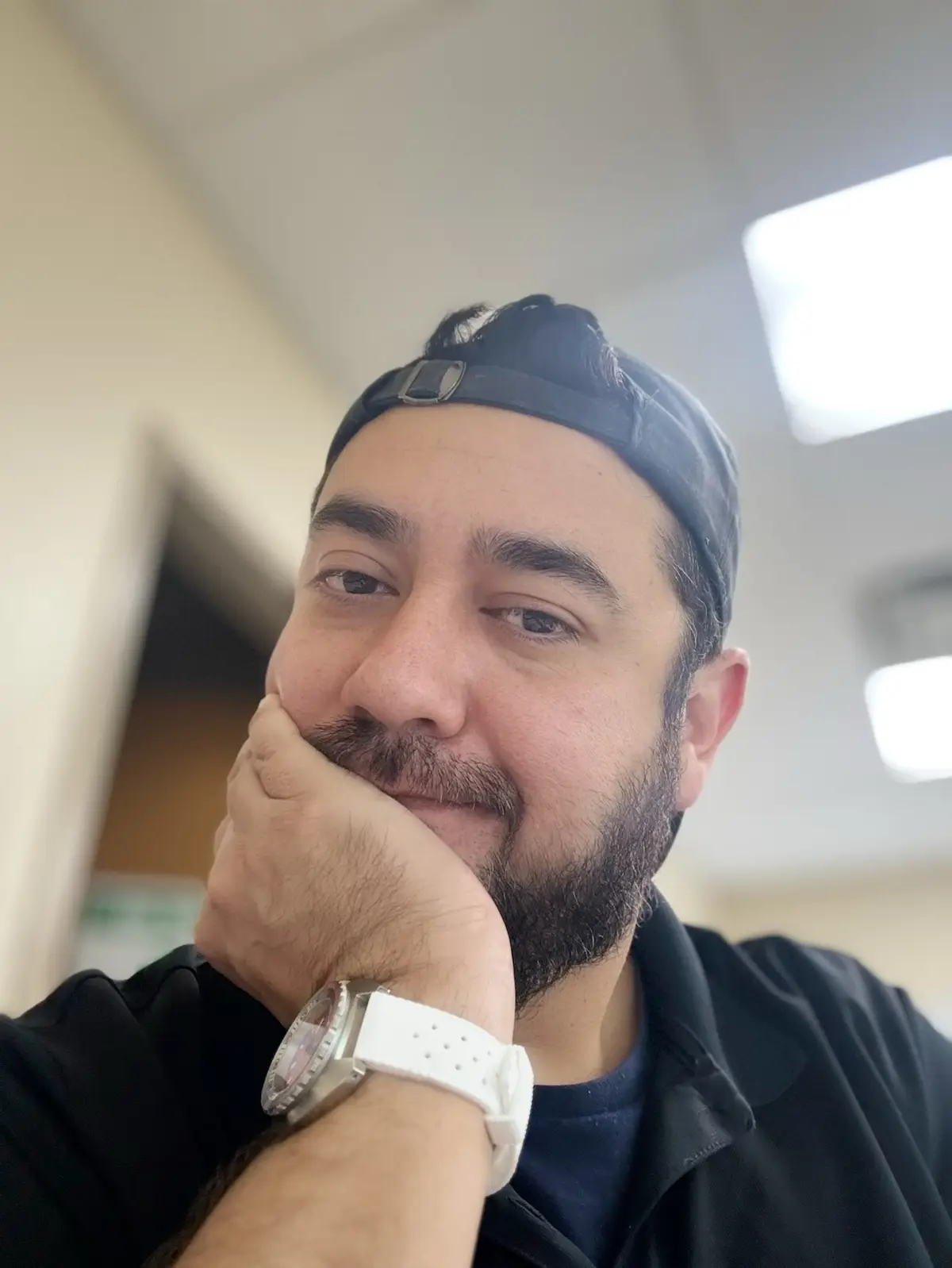 A man with a beard and black cap worn backward poses with his hand on his chin, wearing a white watch, looking thoughtful…