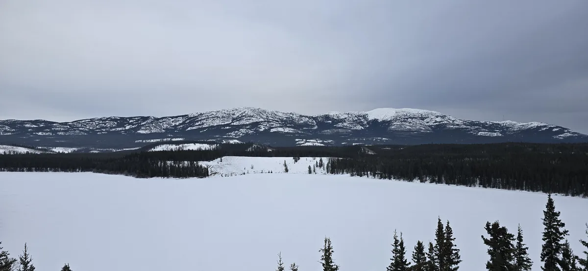 A wide, snowy landscape featuring a frozen lake in the foreground, evergreen trees along the shoreline, and snow-dusted…