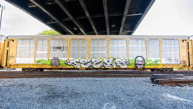 A yellow CSX freight train car sits under a bridge, covered in graffiti including a large, stylized 'JAWS' in white and…