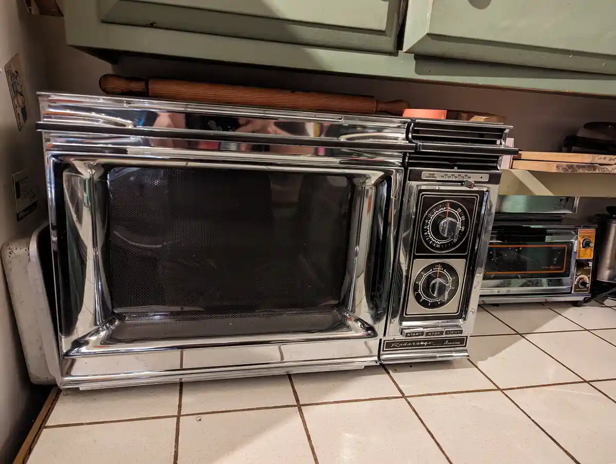 A vintage chrome Amana Radarange microwave oven sits on a tiled kitchen floor, its dials and controls visible, with a wooden…