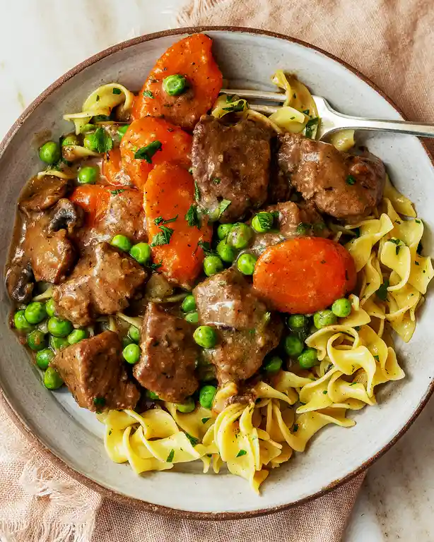 A bowl of beef stew with carrots, peas, mushrooms, and egg noodles, garnished with parsley and served with a fork. The…