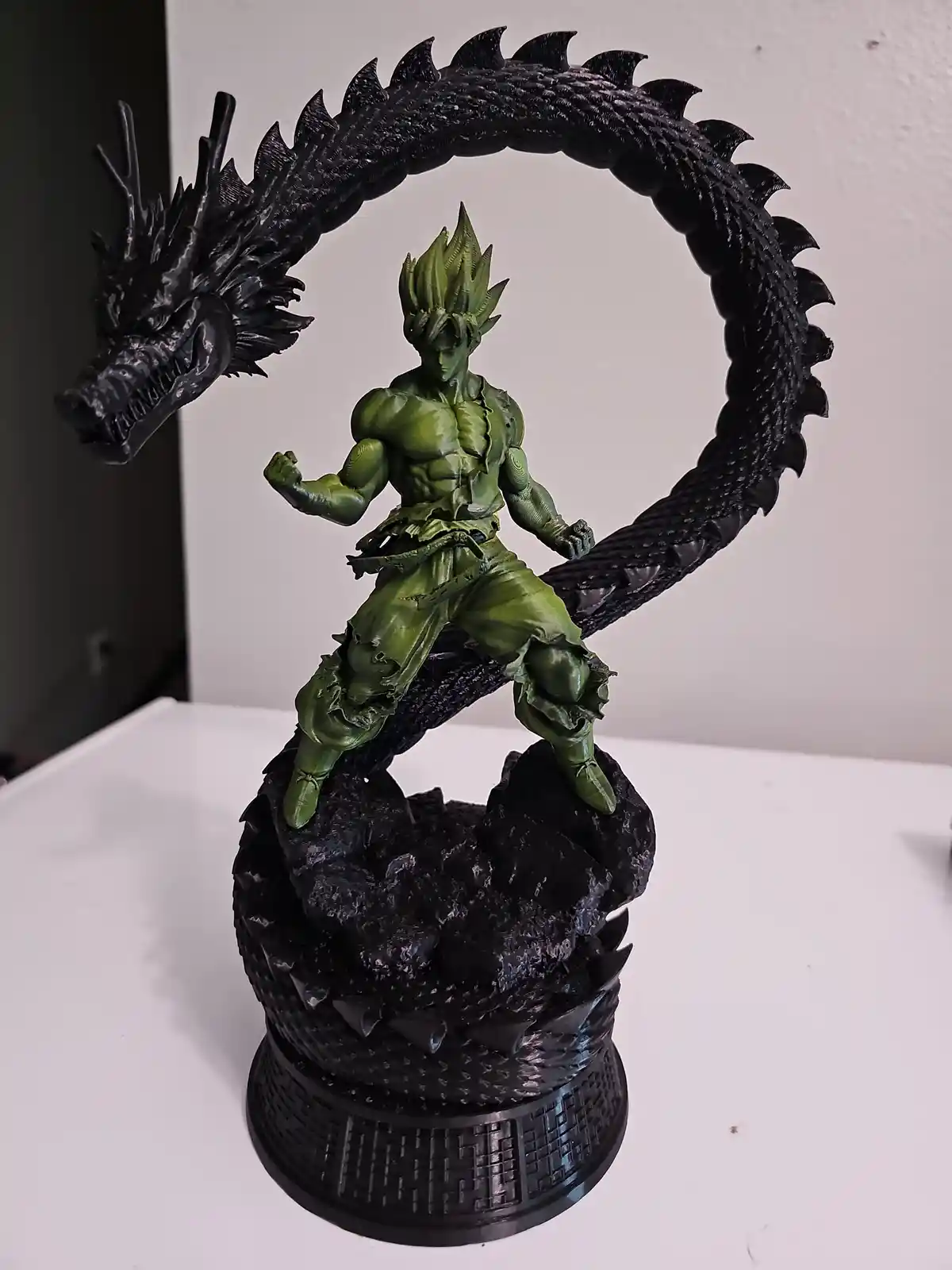 A green, muscular figurine of a character with spiky hair stands atop a black rocky base, encircled by a large black dragon…