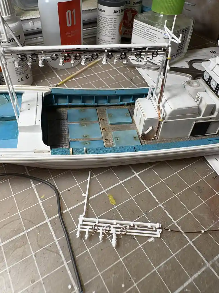 A close-up photo shows a detailed model boat under construction on a tiled work surface, with various paint bottles and…