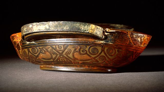 a close-up photograph of an ancient, ornate bowl with a translucent amber-colored body and intricate dark brown geometric…