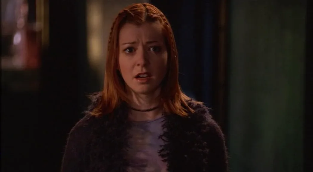 a woman with red hair and braids wears a dark fuzzy cardigan over a purple shirt, looking shocked with wide eyes and an open…