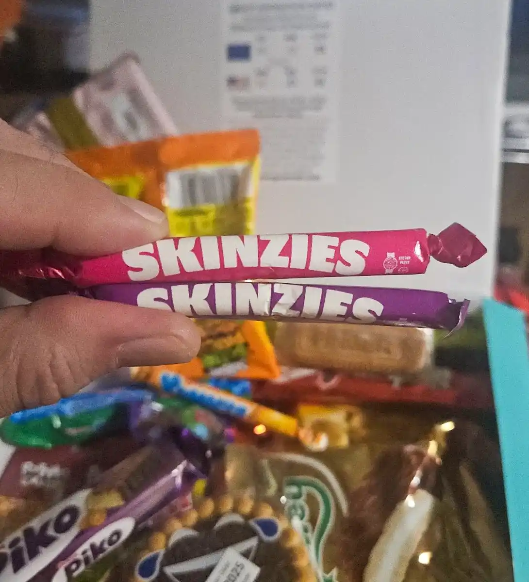 A hand holds two Skinzies candy bars, one pink and one purple, both with the brand name visible. The background is a blurry…