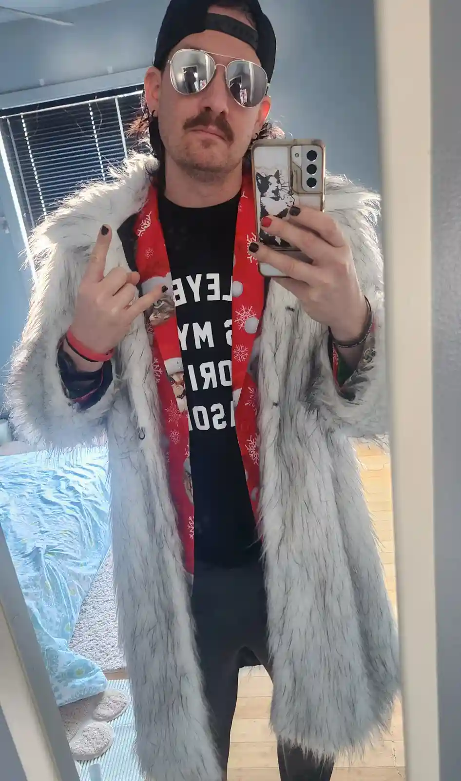 A person takes a mirror selfie wearing a fluffy white coat, a red Christmas-themed scarf with cat images, and a black…