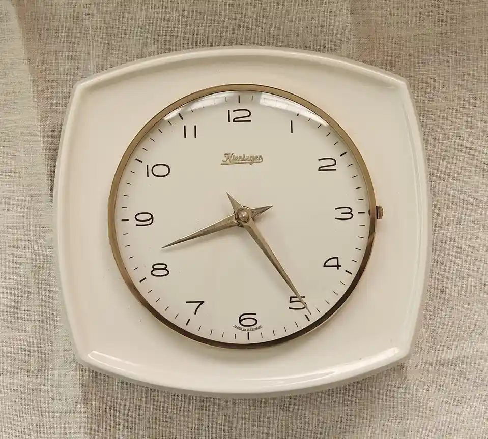 A vintage white Kieninger wall clock with gold trim and black numerals shows the hands pointing to 5:00, but the overlaid…