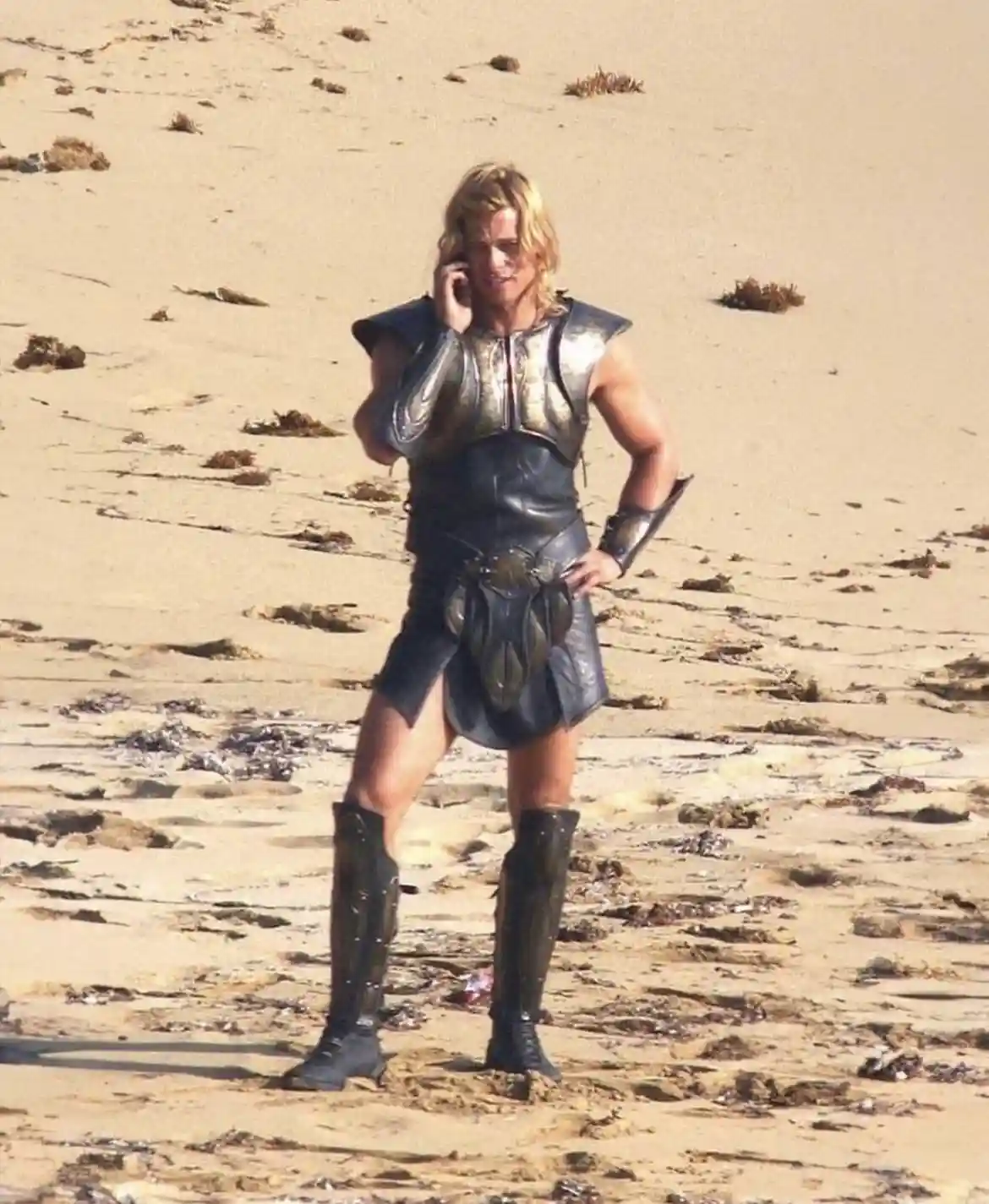 A man in ancient warrior armor stands on a sandy beach, holding a modern cell phone to his ear with one hand while resting…
