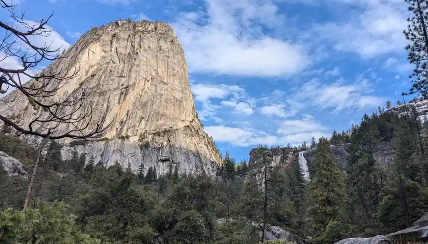 A scenic photo of El Capitan in Yosemite National Park, a massive granite monolith under a partly cloudy blue sky, framed by…