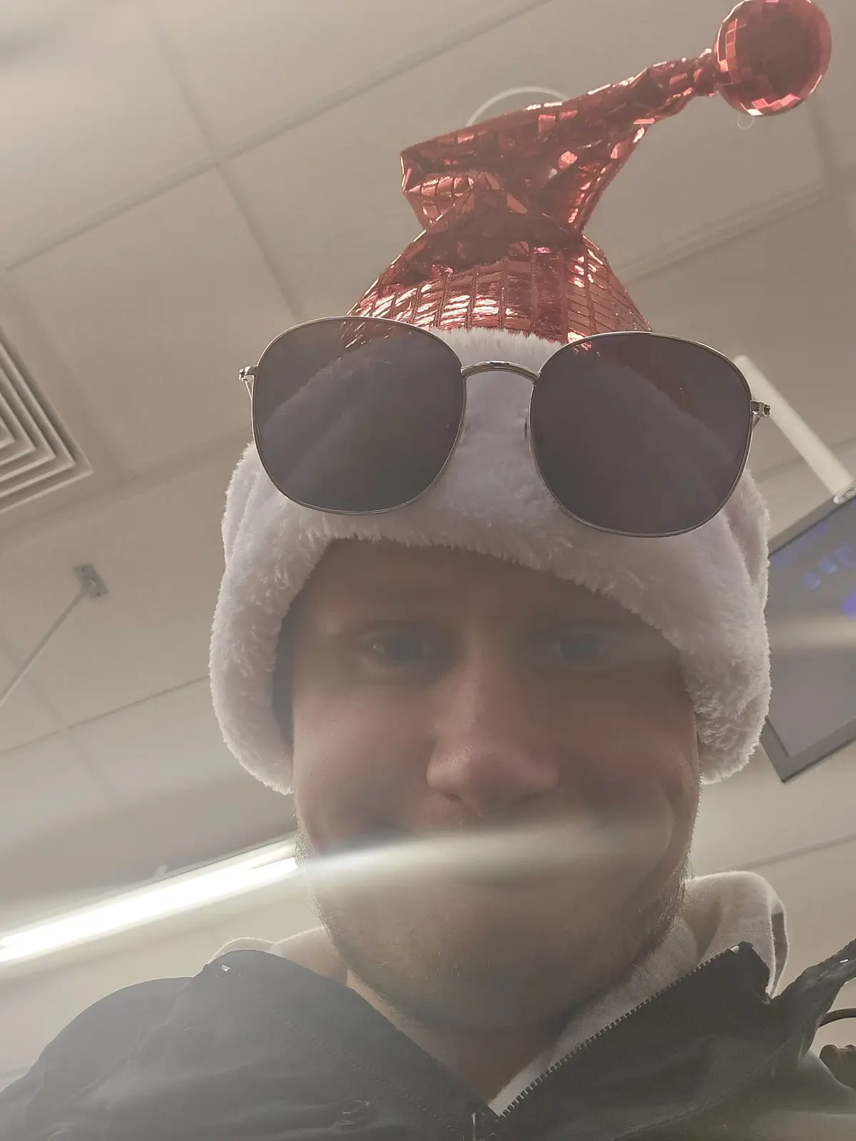 A person wearing a white fuzzy Santa hat with a shiny pink metallic cone and a red disco ball-like pom-pom on top, with a…