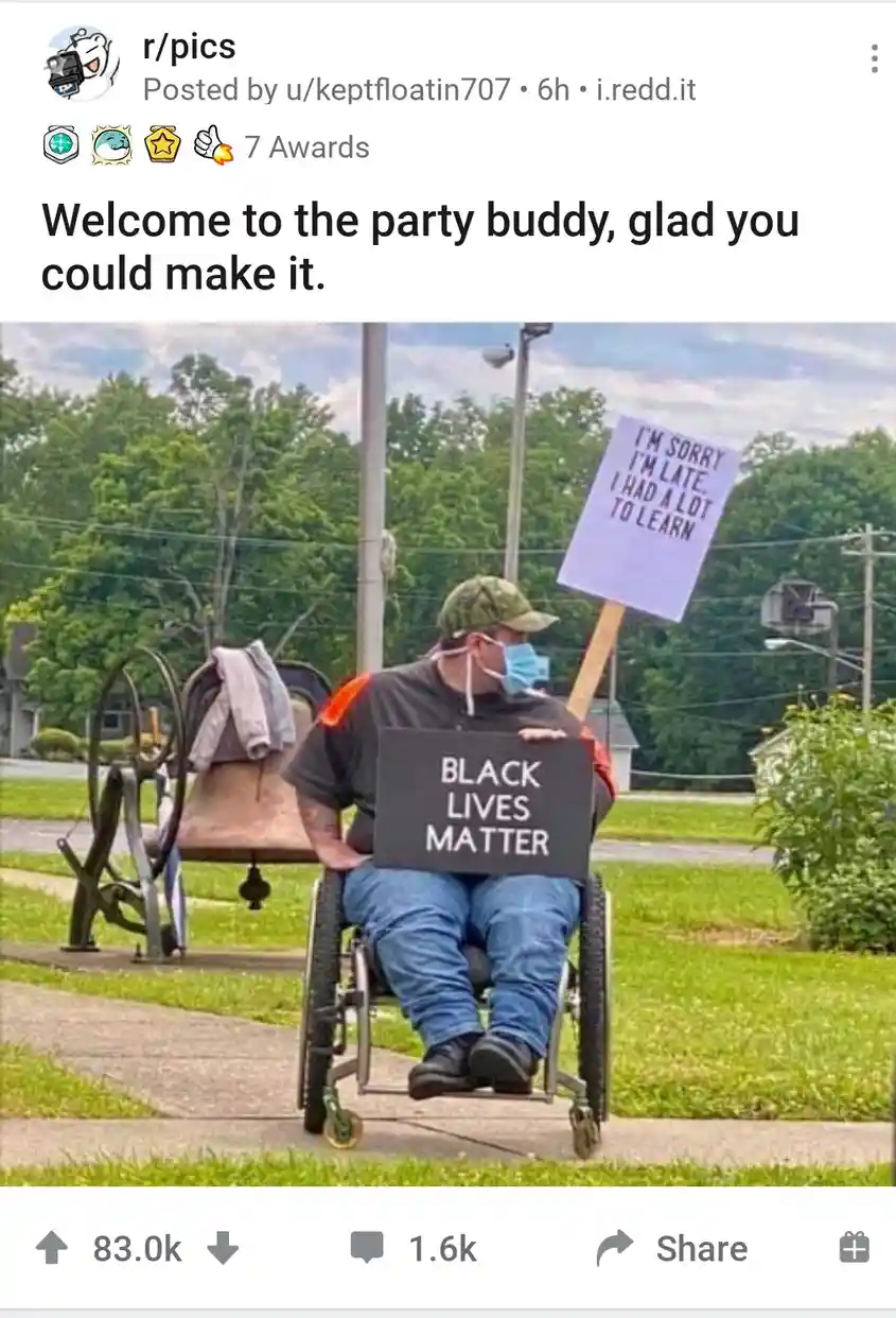 A man in a wheelchair wearing a face mask and a camouflage cap holds two protest signs: one reads 'BLACK LIVES MATTER' and…
