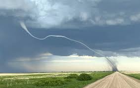 A dramatic photograph shows a long, winding tornado funnel stretching across a rural landscape, appearing to connect a…