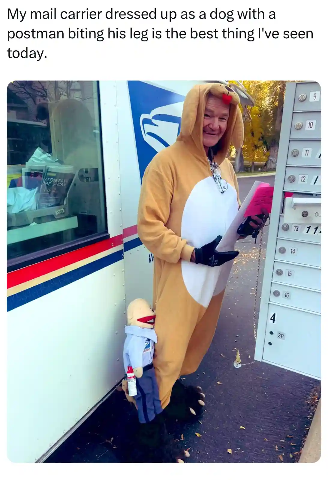 A smiling mail carrier in a tan and white dog costume stands next to a USPS truck and a row of mailboxes. A small puppet…