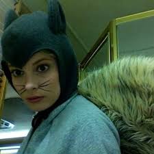 A person wearing a dark green cat-eared hood with whisker markings on their cheeks, looking directly at the camera with a…