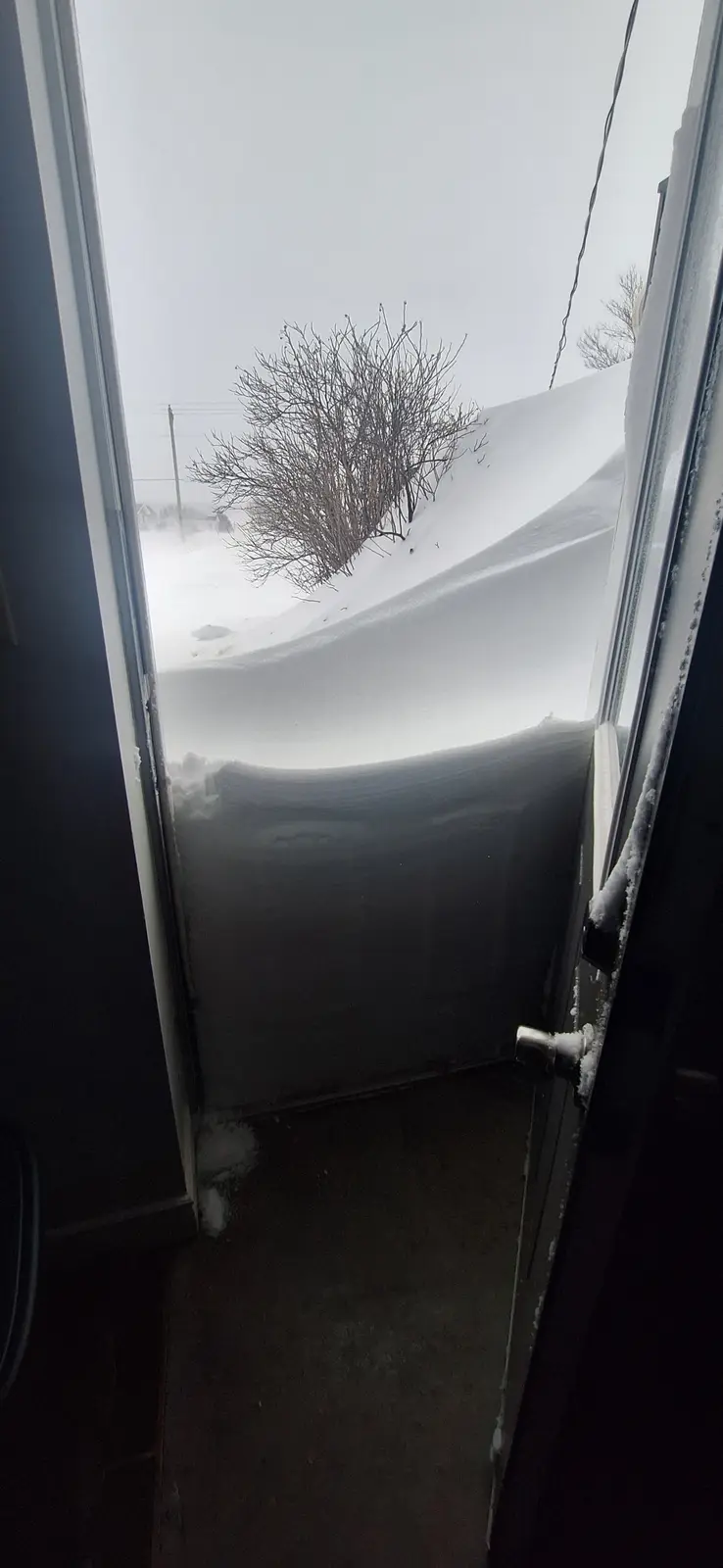 A view from inside a house looking out through a doorway at a massive, wind-blown snowdrift that completely blocks the…