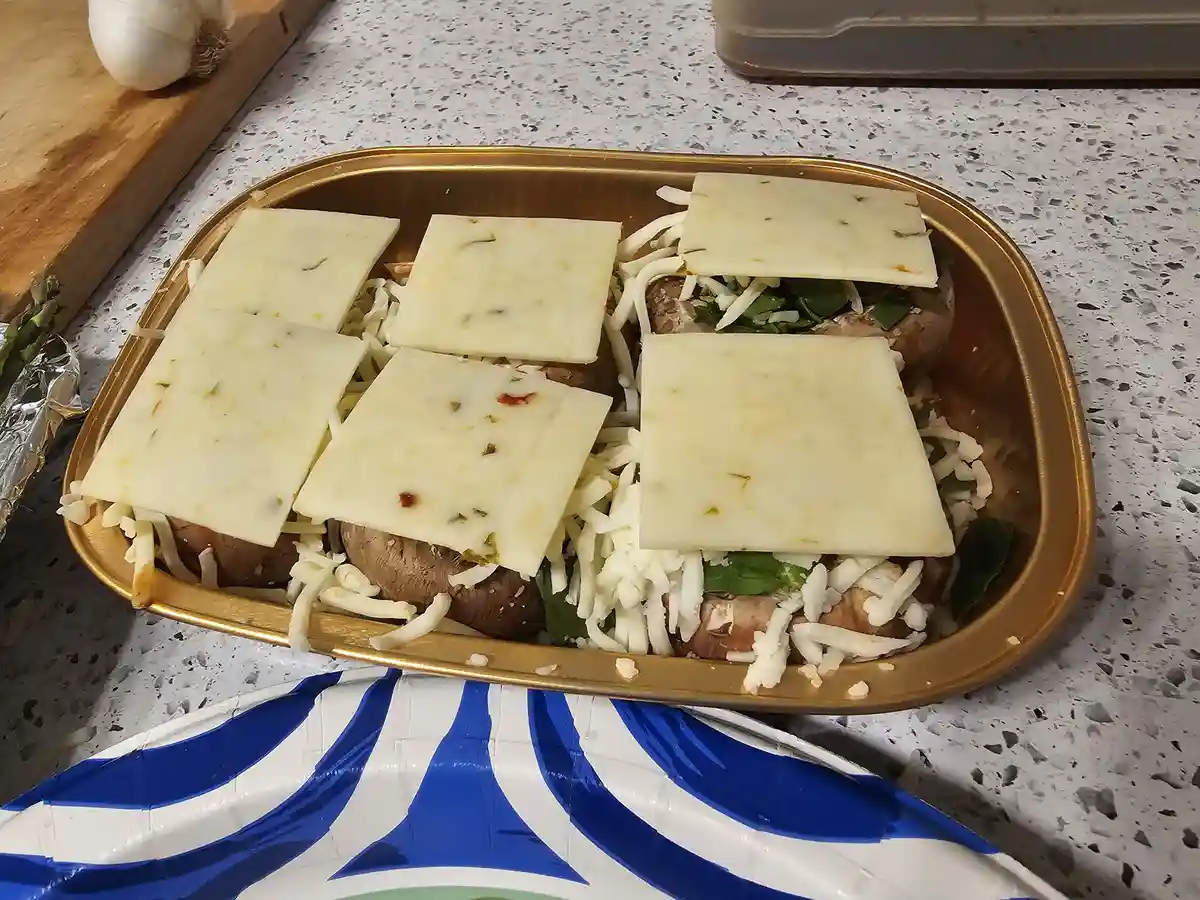 A golden baking tray holds several stuffed mushrooms topped with shredded cheese and square cheese slices. The mushrooms are…