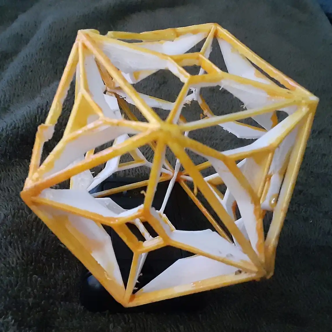 A 3D-printed, geometric twenty-sided die (d20) with its edges painted yellow and white paint dripping down its sides…
