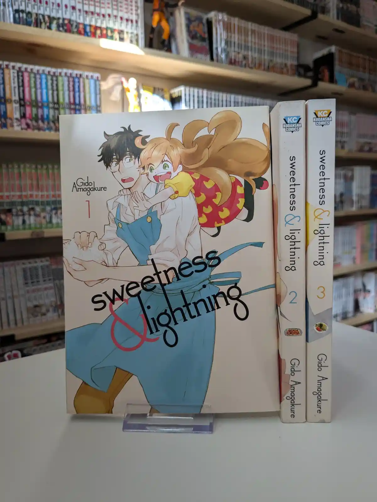 Three volumes of the manga 'Sweetness & Lightning' by Gido Amagakure are displayed on a shelf in a bookstore, with volume 1…