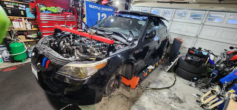 A black BMW car is shown in a cluttered garage with its hood open, engine exposed, and front bumper removed. A red engine…