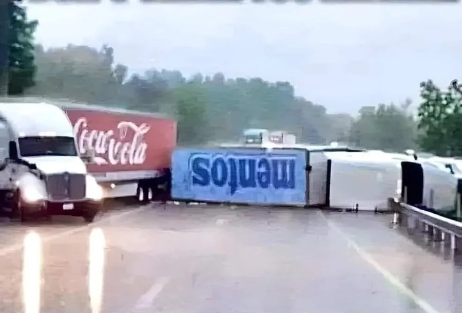 A blurry, low-resolution image shows a white Coca-Cola semi-truck on a wet highway, seemingly about to collide with a blue…
