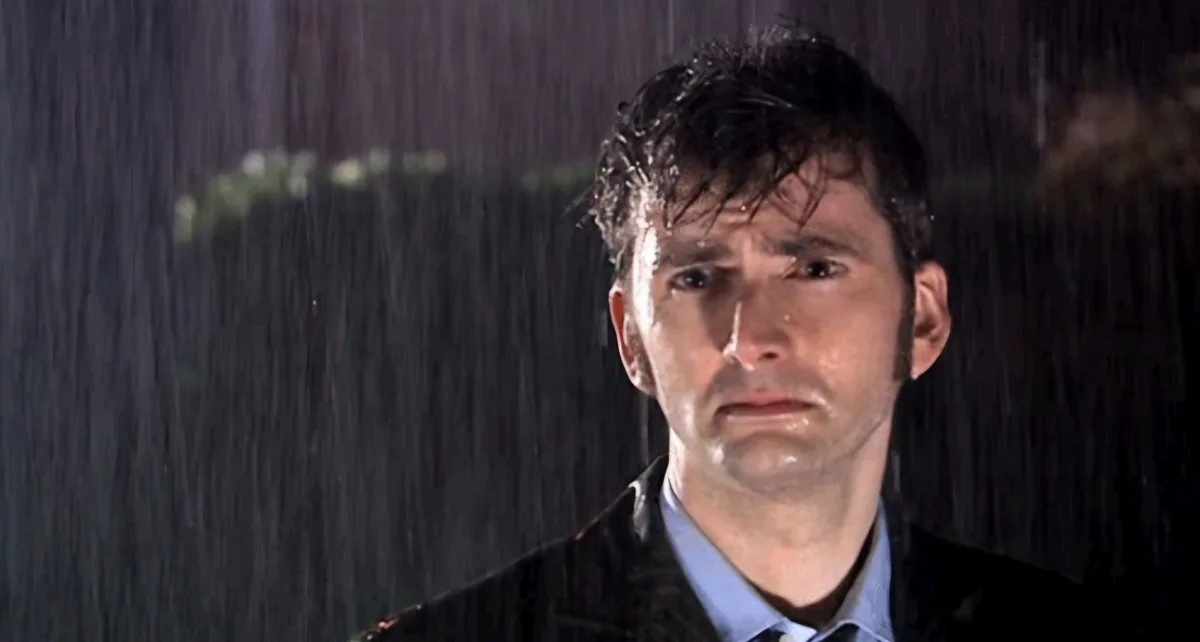 A man with wet hair and a distressed expression stands in heavy rain, wearing a dark coat over a light blue shirt and tie…