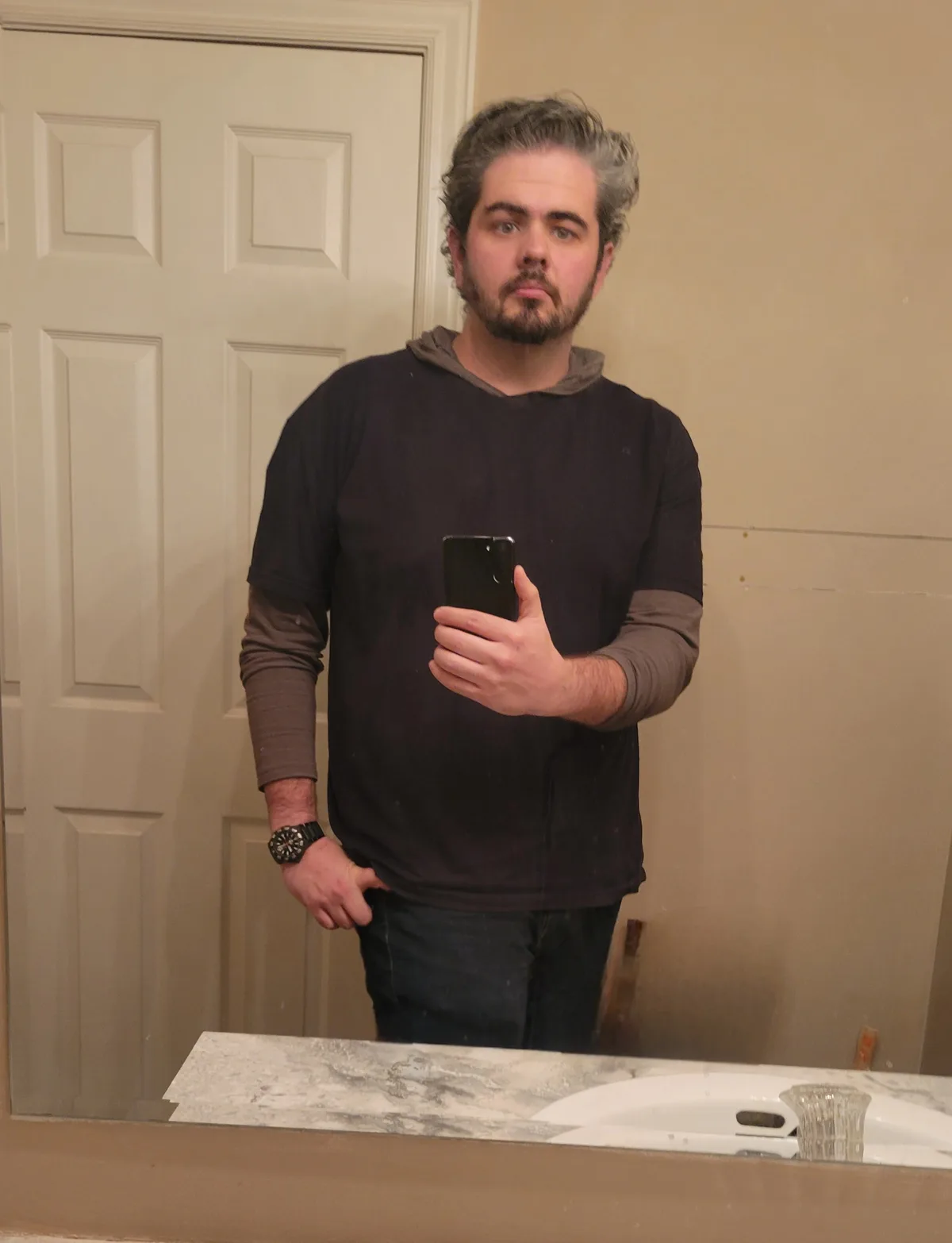 A man with graying hair and a beard takes a mirror selfie in a bathroom, holding a phone in one hand and wearing a dark…