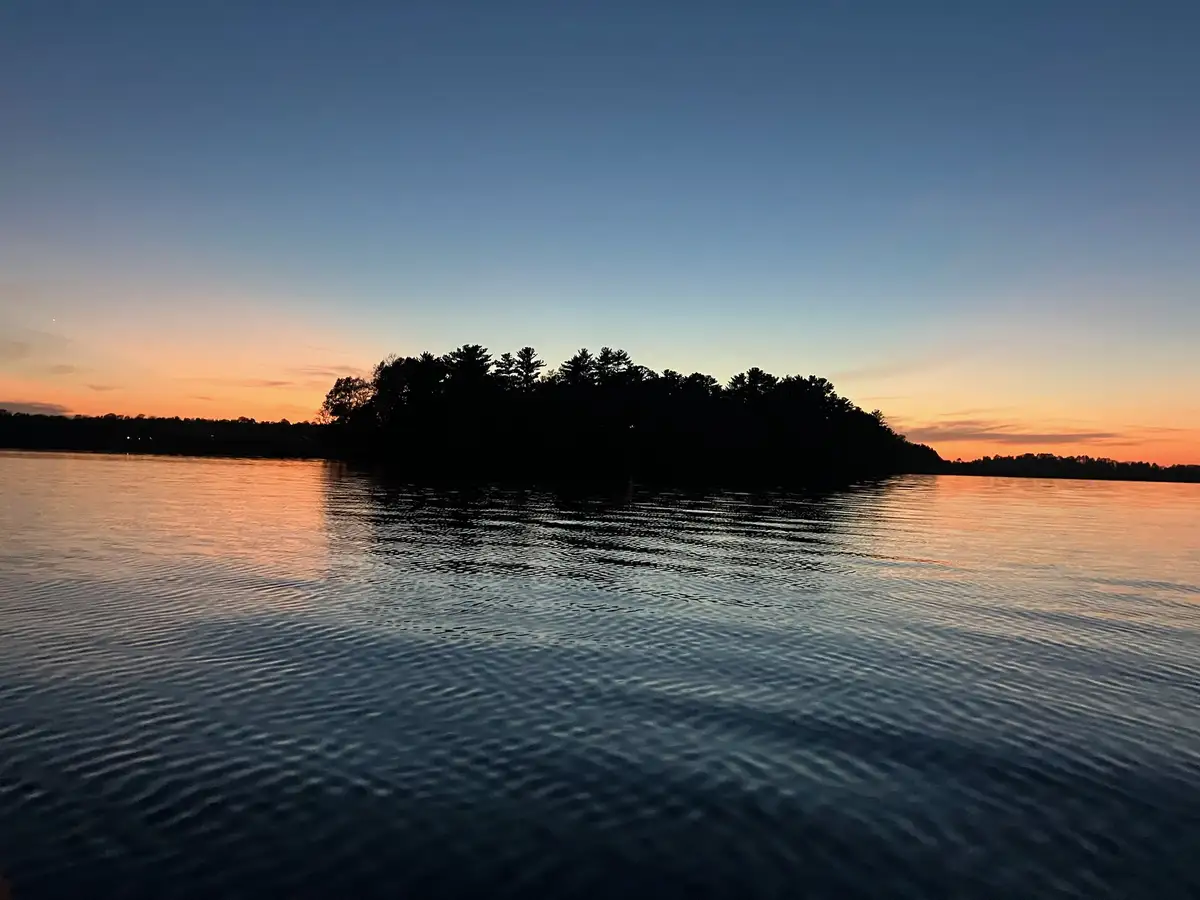 A serene sunset over a calm body of water with a dark tree-lined island in the middle. The sky transitions from orange near…
