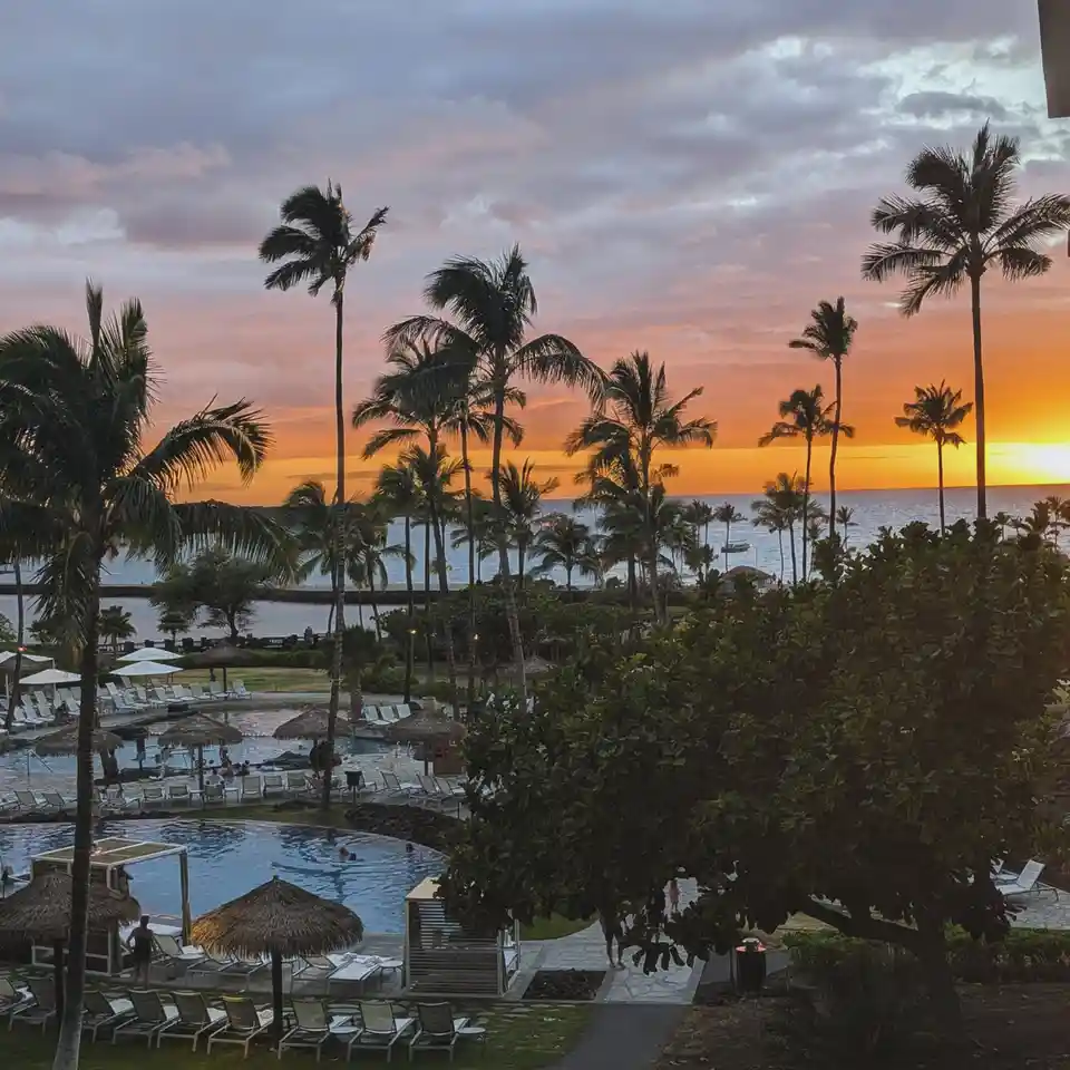 A scenic tropical resort at sunset, featuring palm trees, multiple swimming pools with lounge chairs, thatched umbrellas…